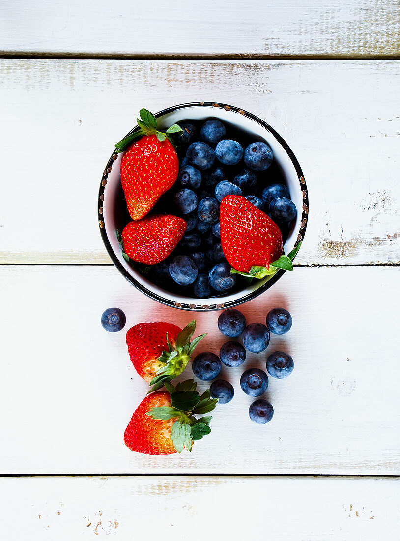 Blueberries and strawberries in a bowl and on a wooden surface (seen from above)