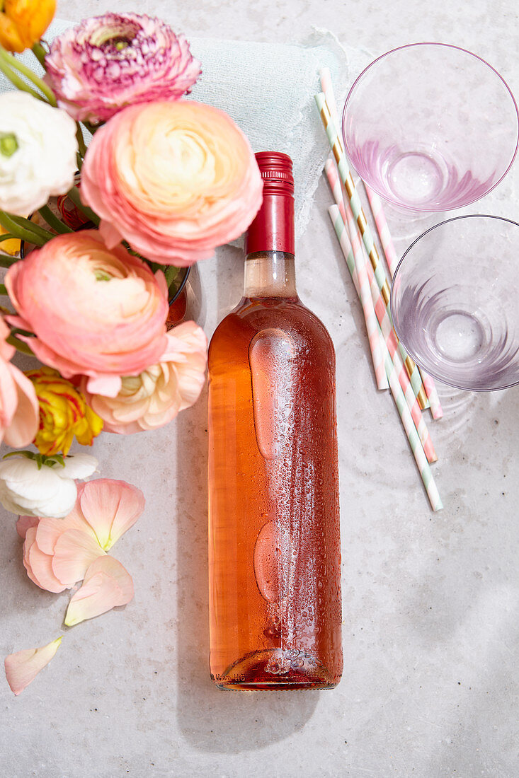 Eine Flasche Roséwein mit Wassertropfen, Strohhalme und Gläser neben Ranunkelstrauß