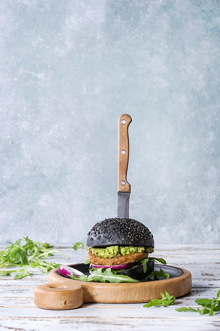 Homemade burger in black bun with avocado, arugula, onion on wood serving board