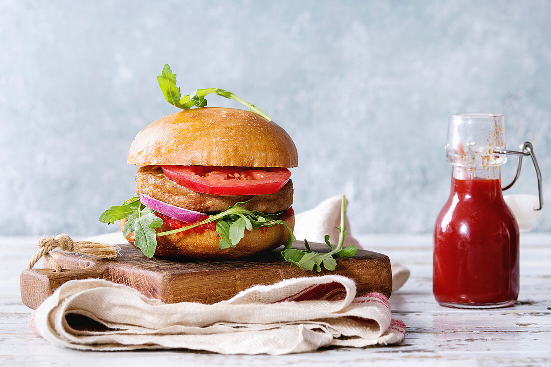 Homemade burger in classic bun with tomato sauce, arugula, meat, cheese, onion, bottle of ketchup on wood serving board