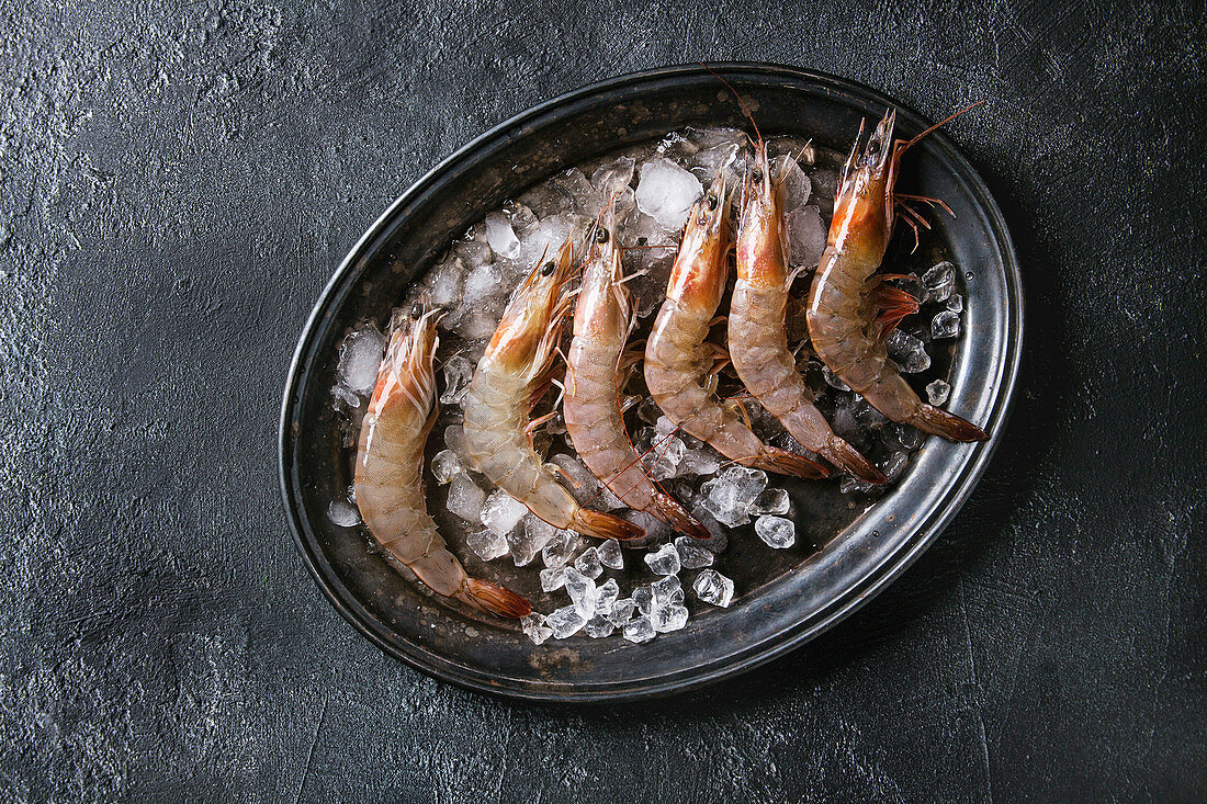 Raw whole fresh uncooked prawns shrimps on ice on vintage metal tray over black texture background
