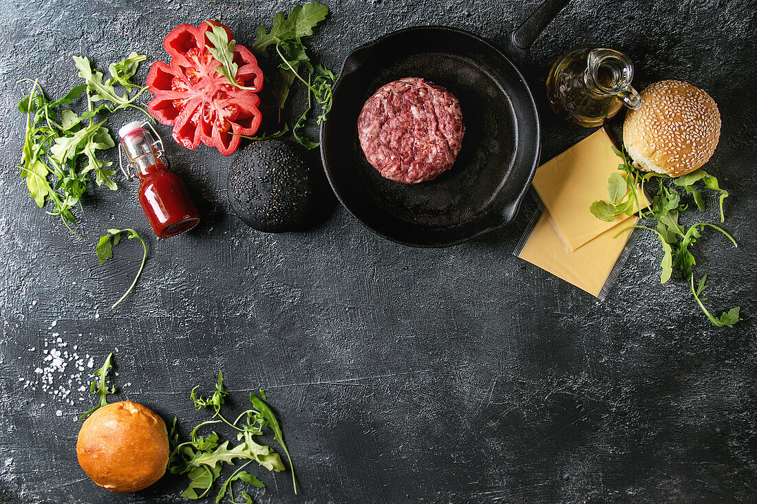 Ingredients for cooking hamburger: Meat beef burger in pan, cheese, ketchup sauce, tomato, black and white buns, arugula salad over dark texture background