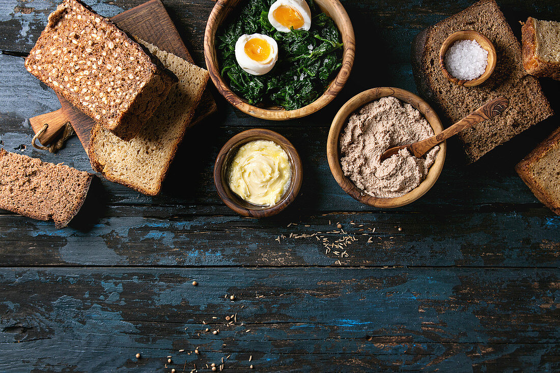 Variety loaves of sliced homemade rye bread whole grain and seeds for breakfast with olive wood bowls of butter, liver paste, salt, spinach, eggs