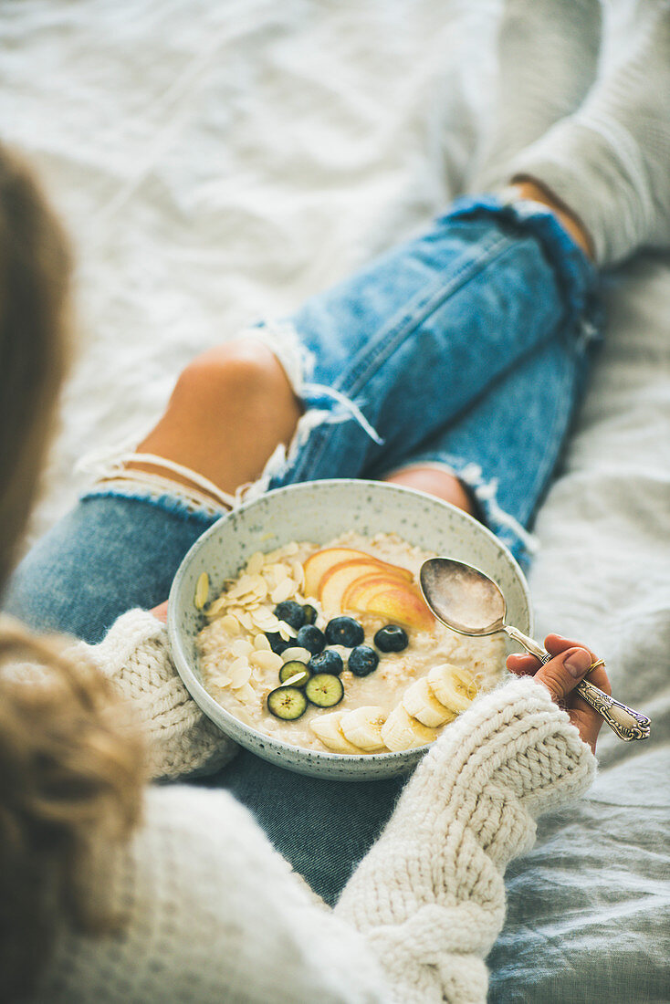 Frau in Wollpullover und Jeans isst veganes Mandeldrink-Porridge mit Früchten