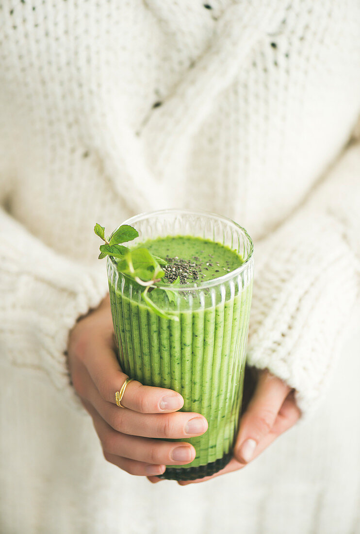 Matcha green vegan smoothie with chia seeds and mint in glass in hands of female wearing white sweater