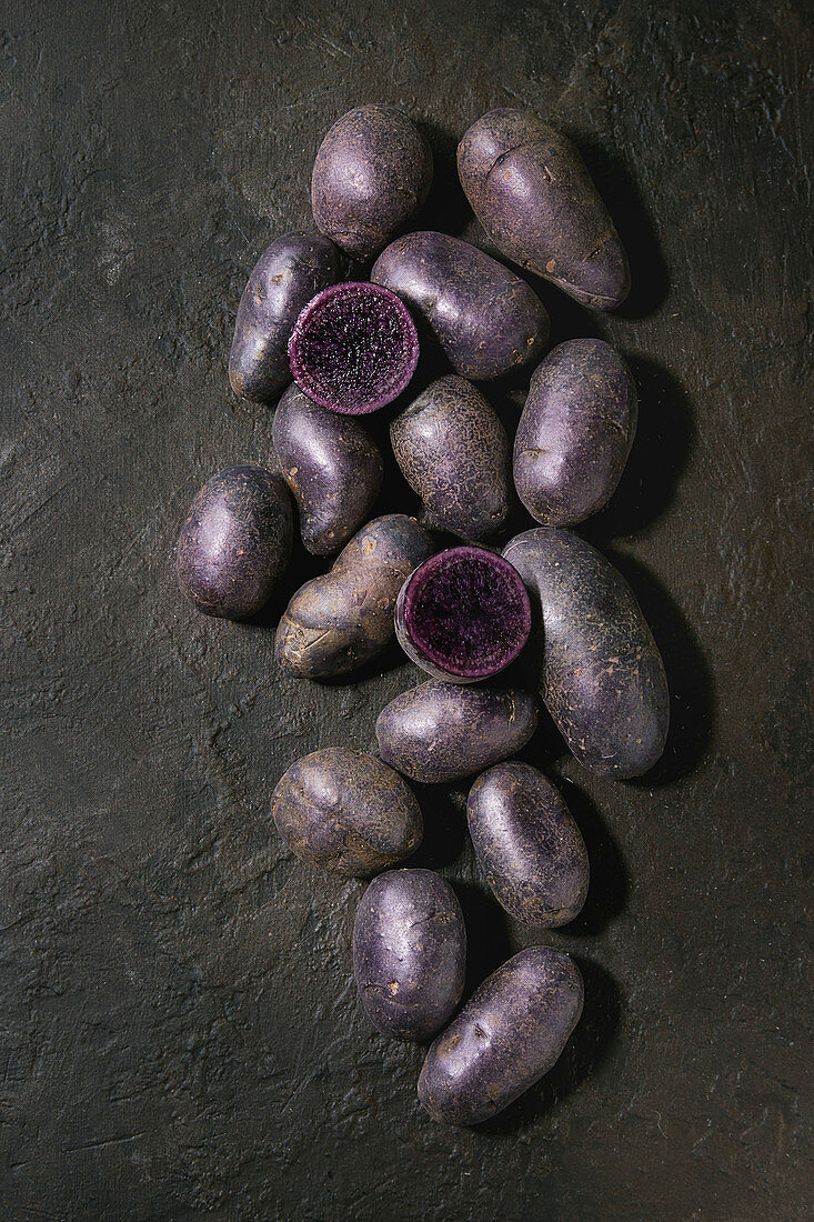 Raw purple uncooked organic potatoes, whole and sliced over dark texture background