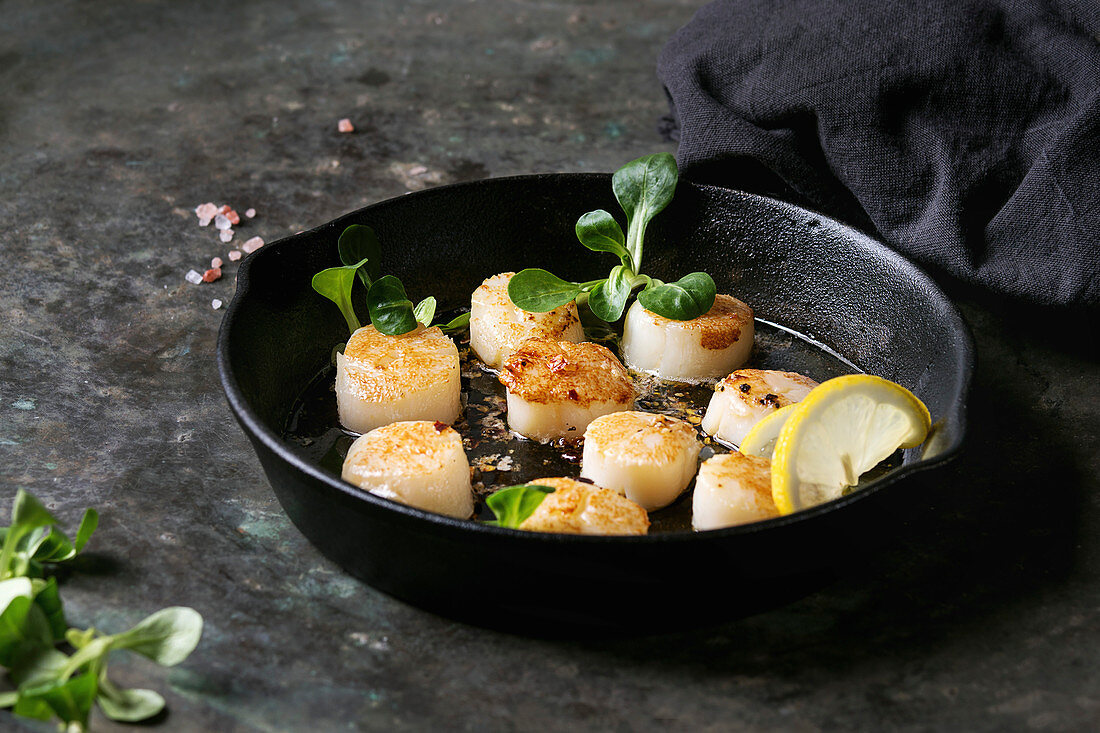 Fried scallops with butter lemon spicy sauce in cast-iron pan served with green salad and textile napkin over old dark metal background