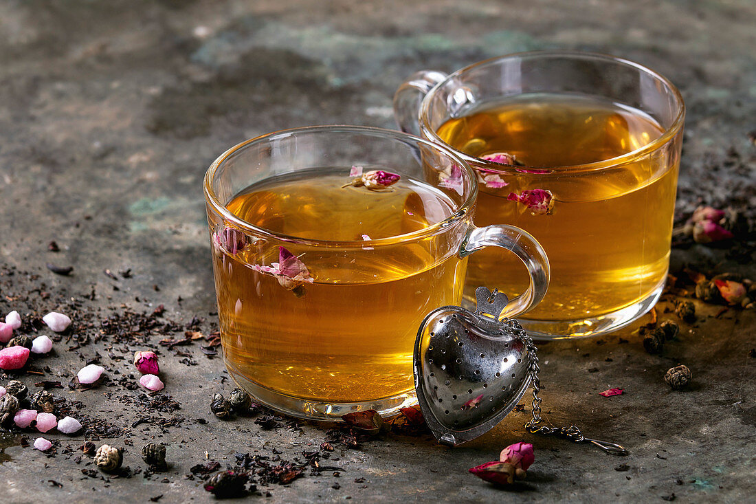 Two glasses of hot tea, rose buds, heart shaped tea strainer, pink sugar over old dark metal background