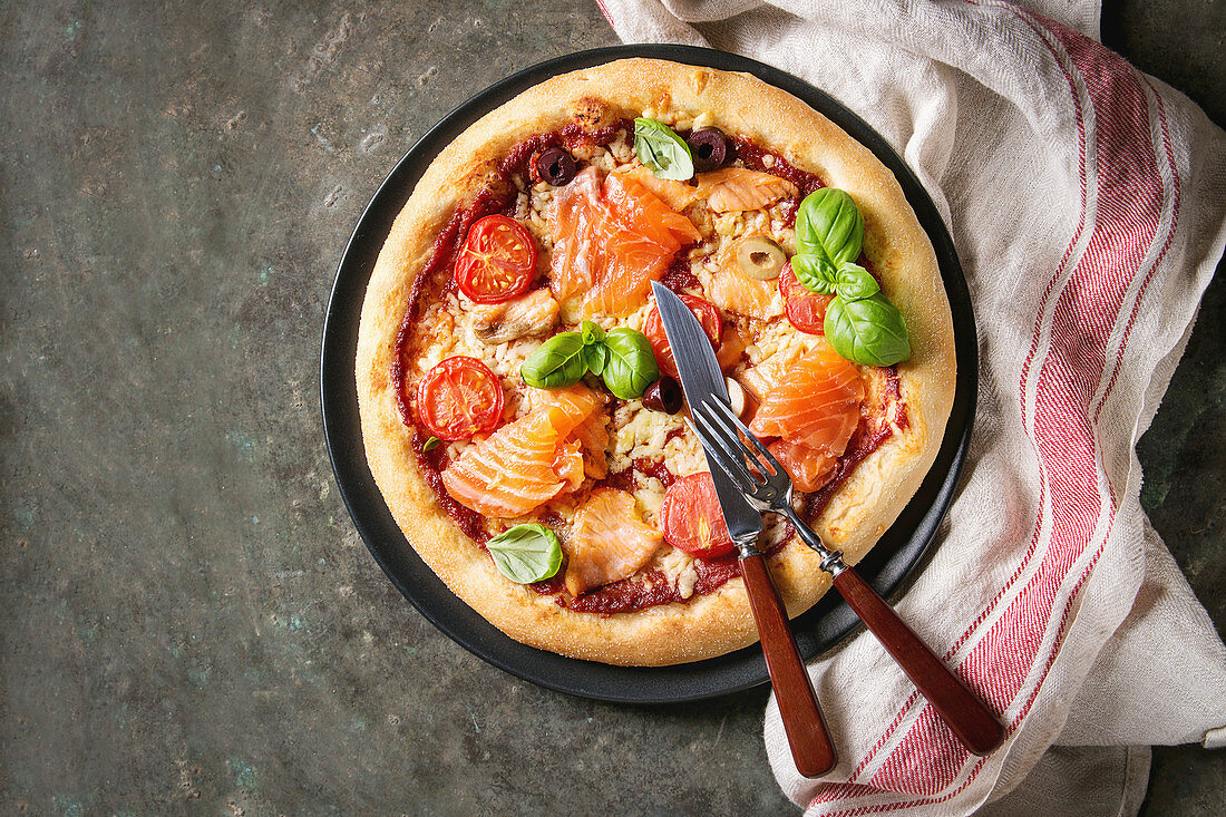 Traditional pizza with smoked salmon, cheese, tomatoes and basil served on black plate with kitchen towel over old dark metal background