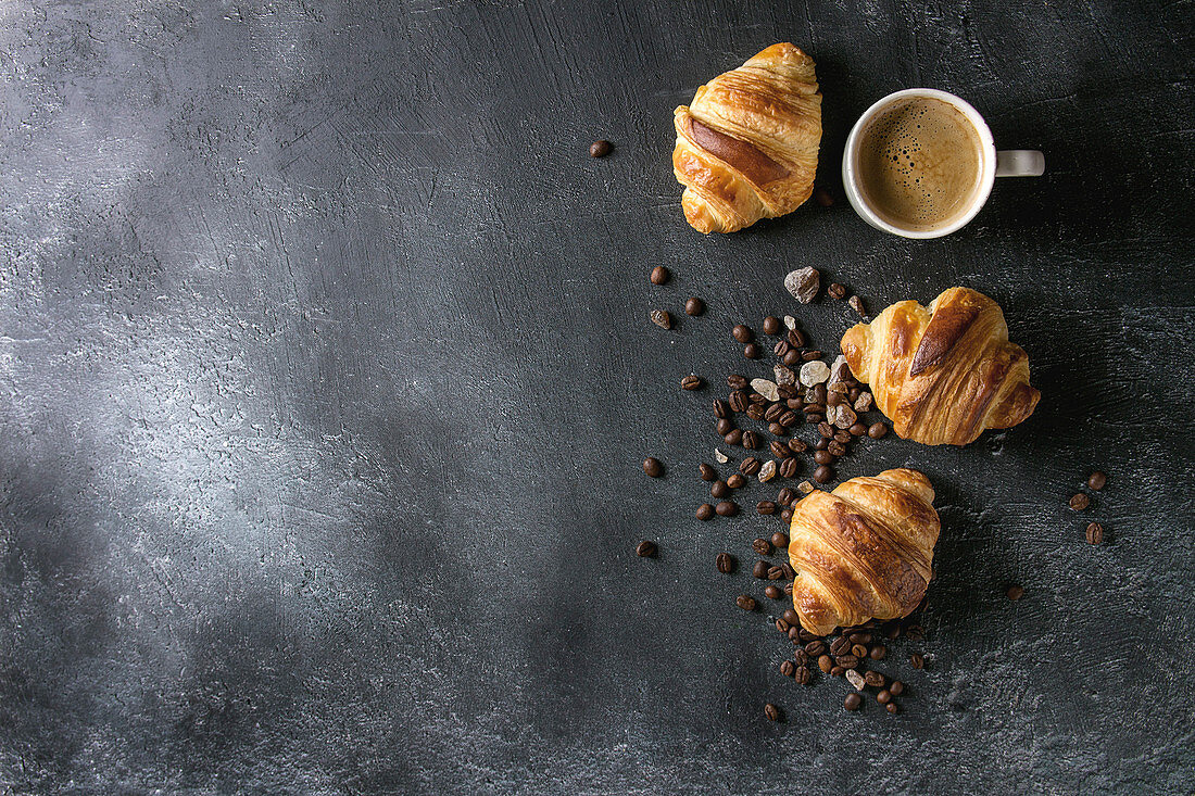 Fresh baked traditional croissants and mug of espresso coffee, coffee beans, sugar over black texture background