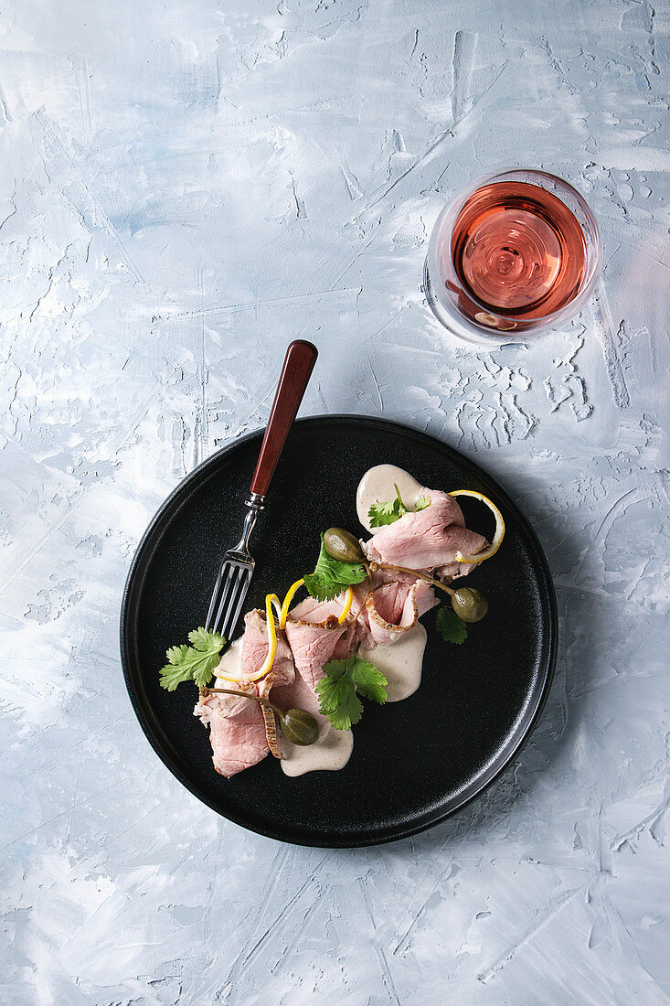 Vitello tonnato italian dish. Thin sliced veal with tuna sauce, capers and coriander served on black plate with fork and glass of rose wine