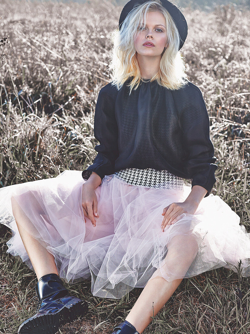 A blonde woman wearing a hat, a black blouse and pink tut sitting in an autumnal meadow