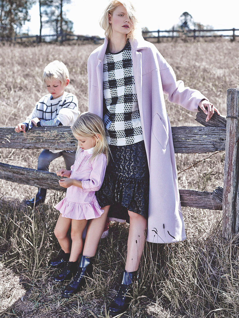 Blonde Frau in schwarz-weiß karierter Bluse, Rock und fliederfarbenem Mantel mit zwei Kindern vor Koppel