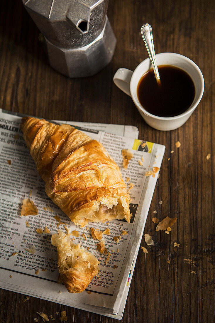 A croissant on newspaper, a coffee pot, and a cup of coffee