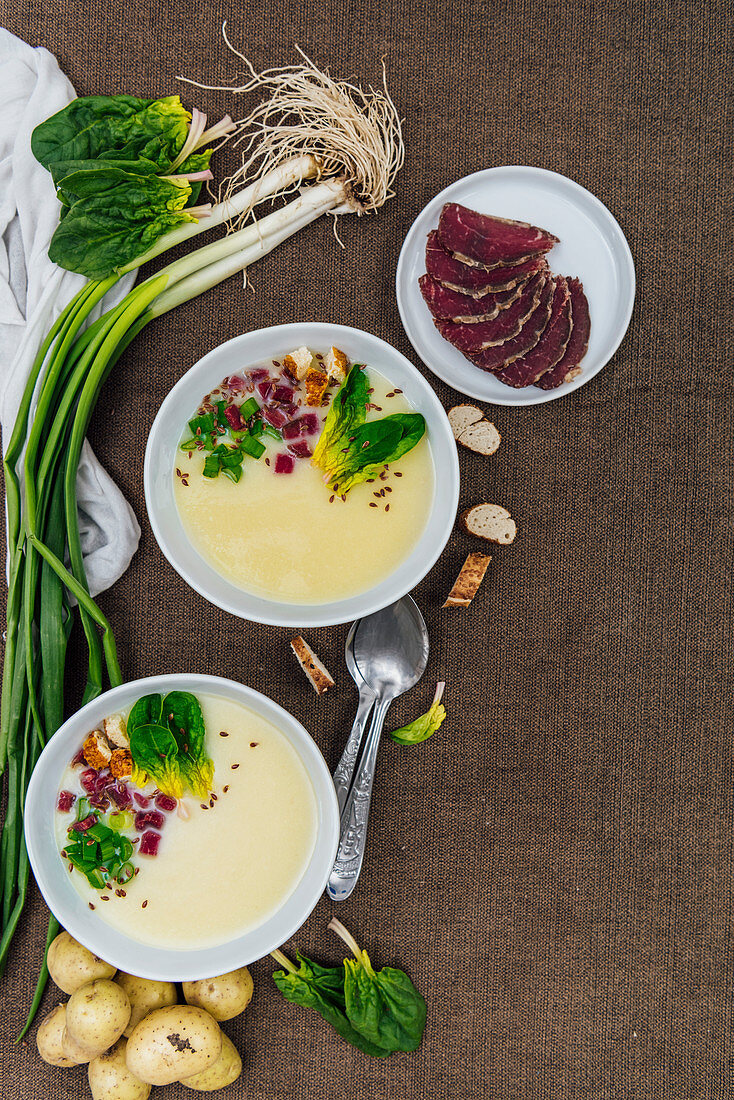 Cream of potato soup topped with green onions, spinach, simit croutons, flax seeds and aged beef in two white bowls