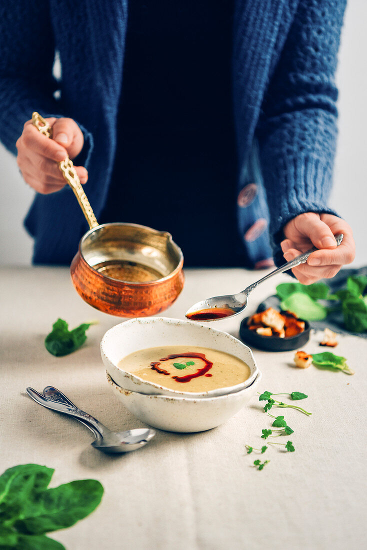 A woman in blue cardigan drizzling a sauce over a bowl of Turkish lentil soup from a copper sauce pan