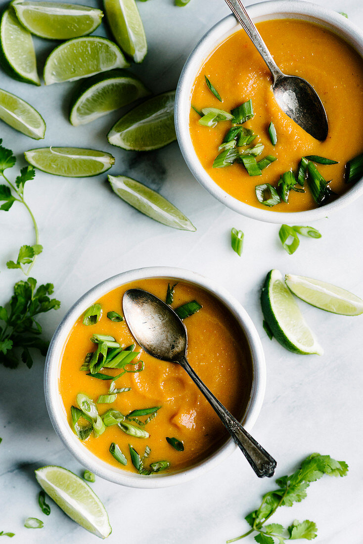 Bowls of butternut squash soup with lime wedges and cilantro on a white marble background