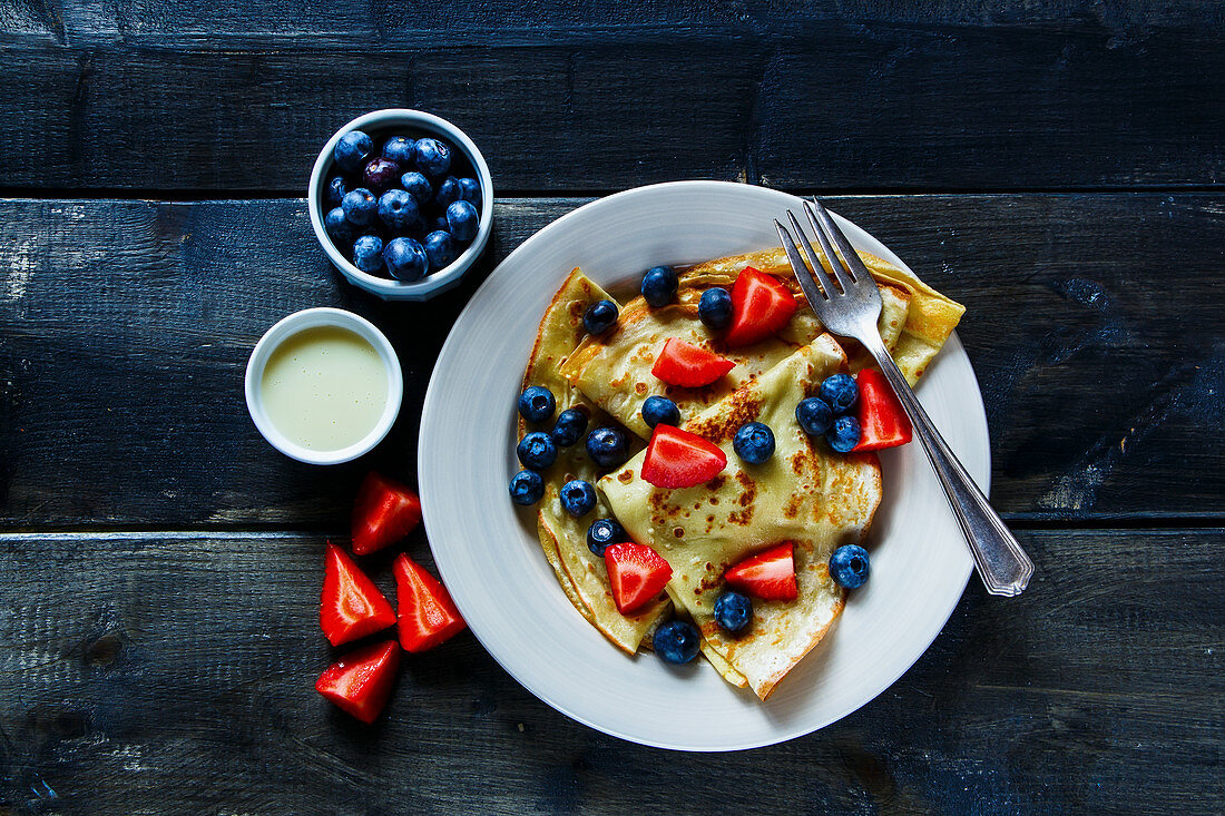 Freshly made thin pancakes or crepes with fresh berries and cream on plate over rustic wooden background