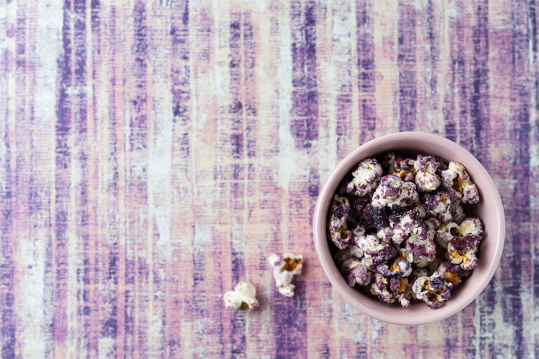 Purple coloured popcorn in a small bowl on a violet surface (top view)