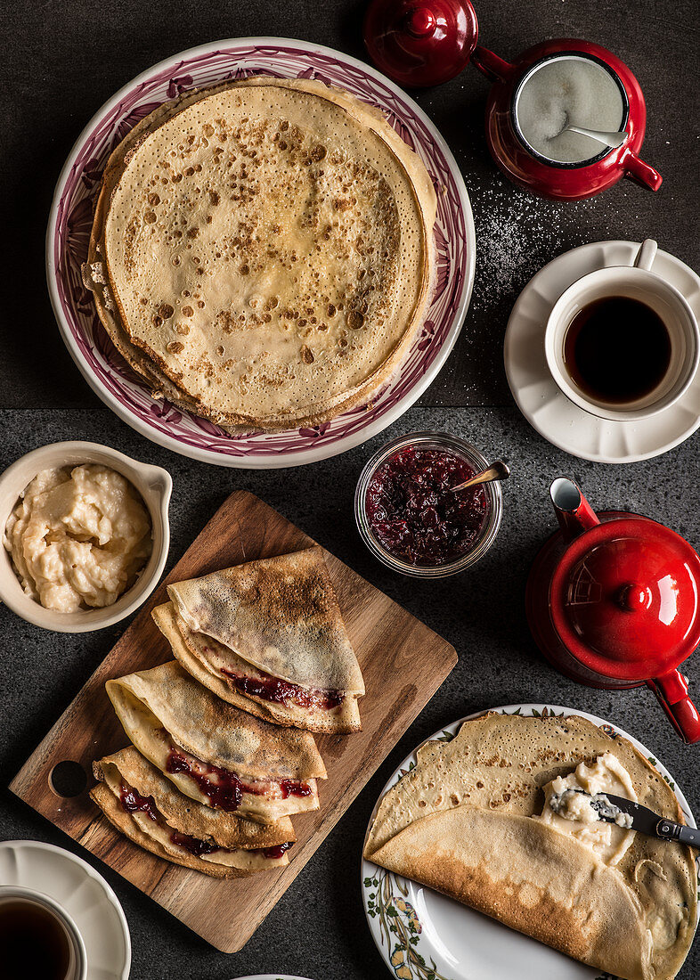 Plates with crepes with jam jar, coffee cup, red coffee pot and red sugar bowl on dark background