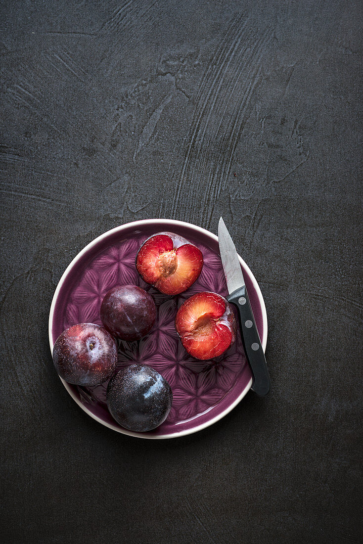 Whole and open red plums with knife on purple plate and dark background