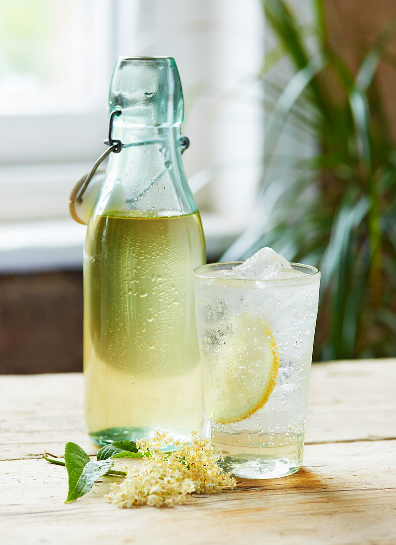 A glass of Elderflower cordial and sparkling water in front of a bottle of Elderflower cordial