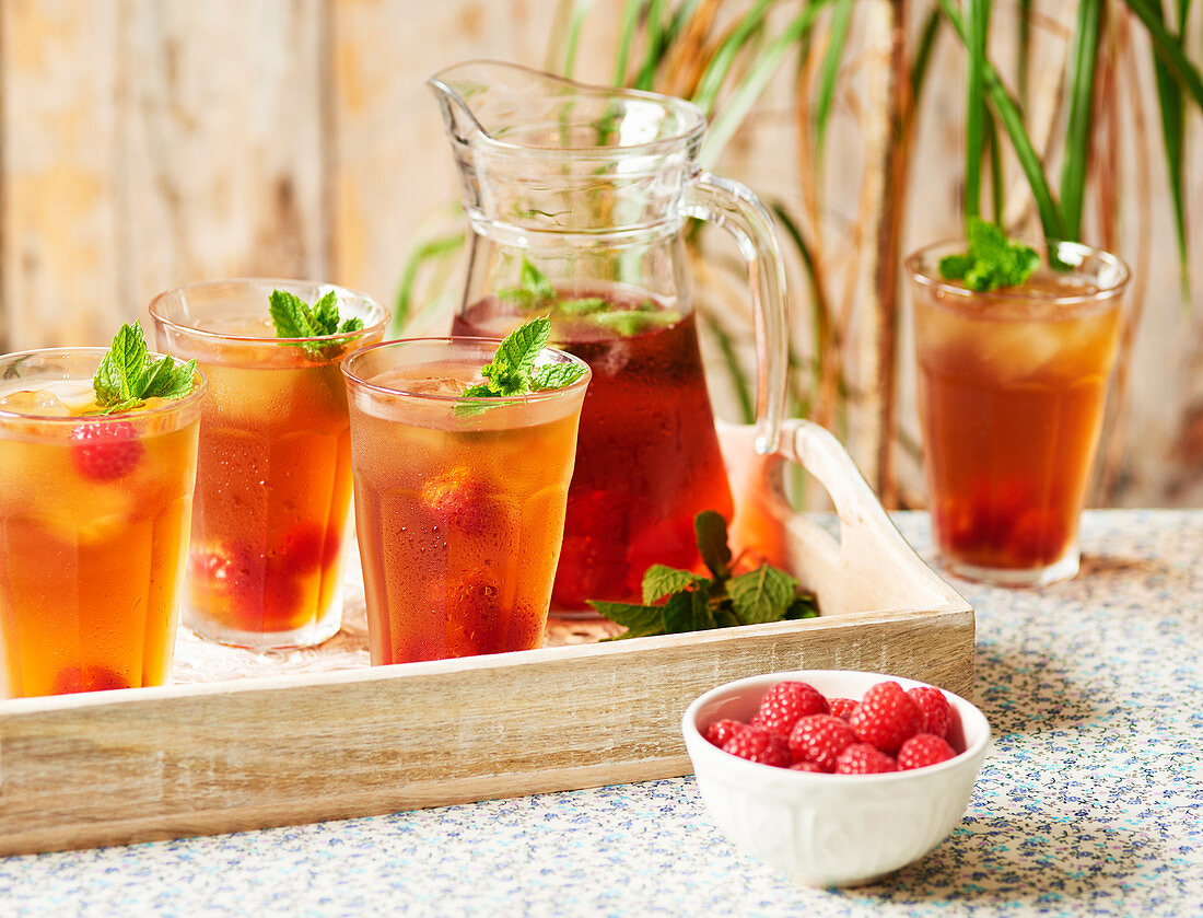 A jug and glasses of Rooibos Iced tea served with raspberies and mint on a wooden tray sitting on a floral table cloth