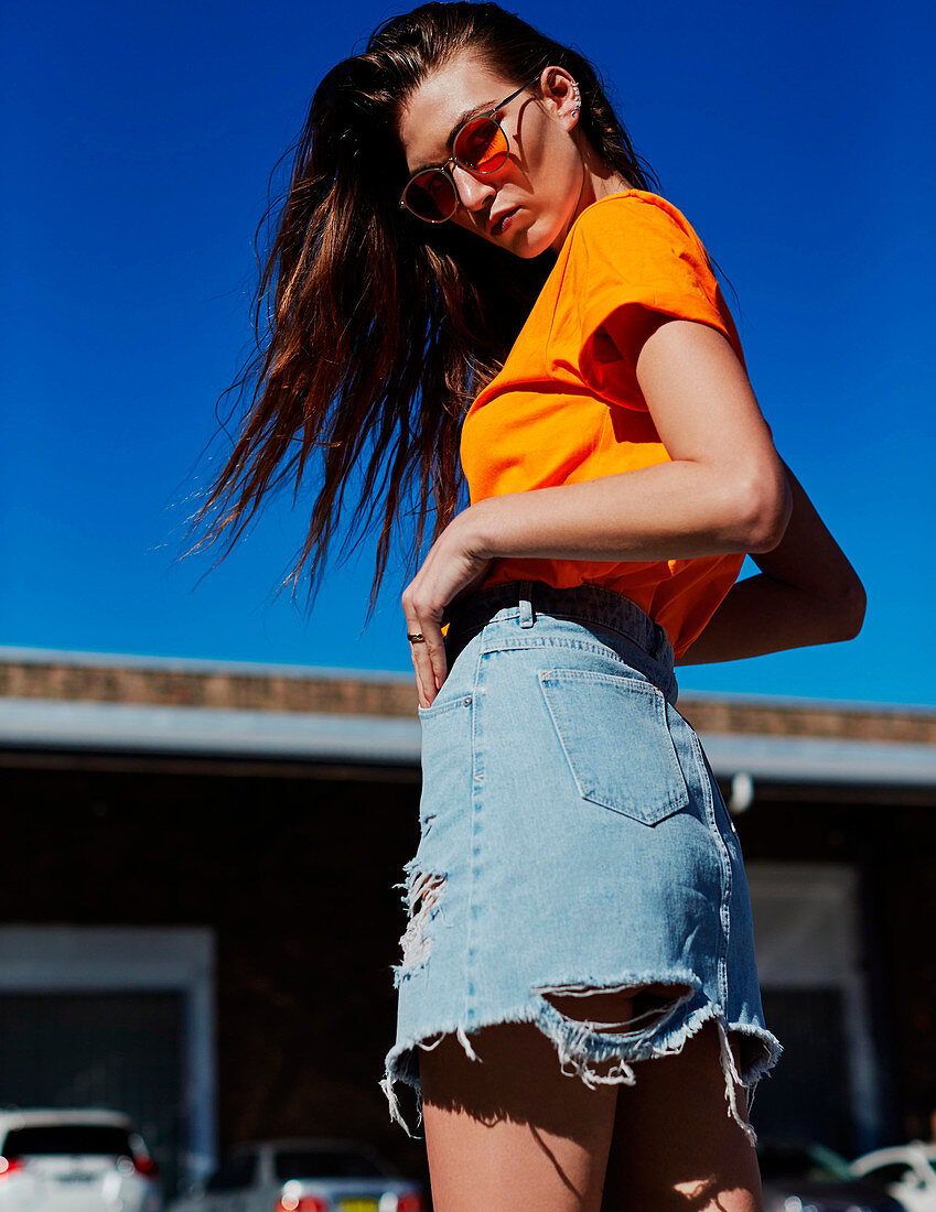 A brunette woman wearing an orange t-shirt and a ripped denim skirt