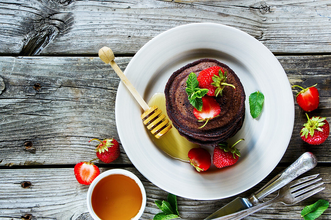 Chocolate pancakes with fresh strawberries and honey, served on ceramic plate over old grey wooden background