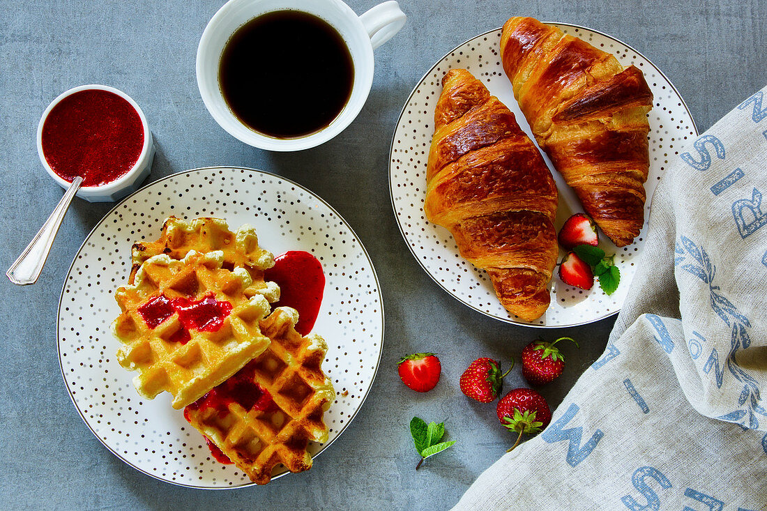 Close up of breakfast table with fresh baked croissants and waffles, served with coffee, strawberries and homemade berry jam