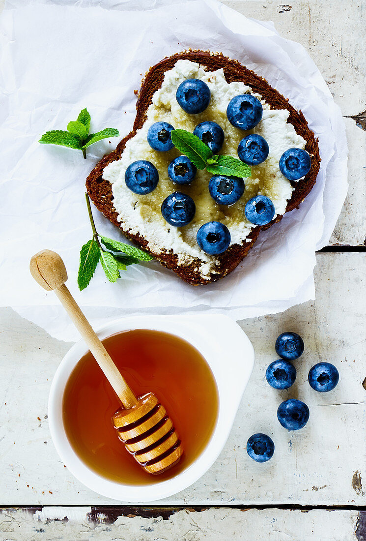 Close up of grunge white wooden table with ricotta, fresh blueberries and honey sandwich on whole grain bread