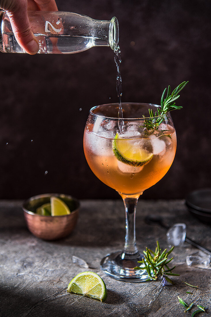 Pink gin cocktail with angostura bitters, lime and rosemary, tonic water beeing poured