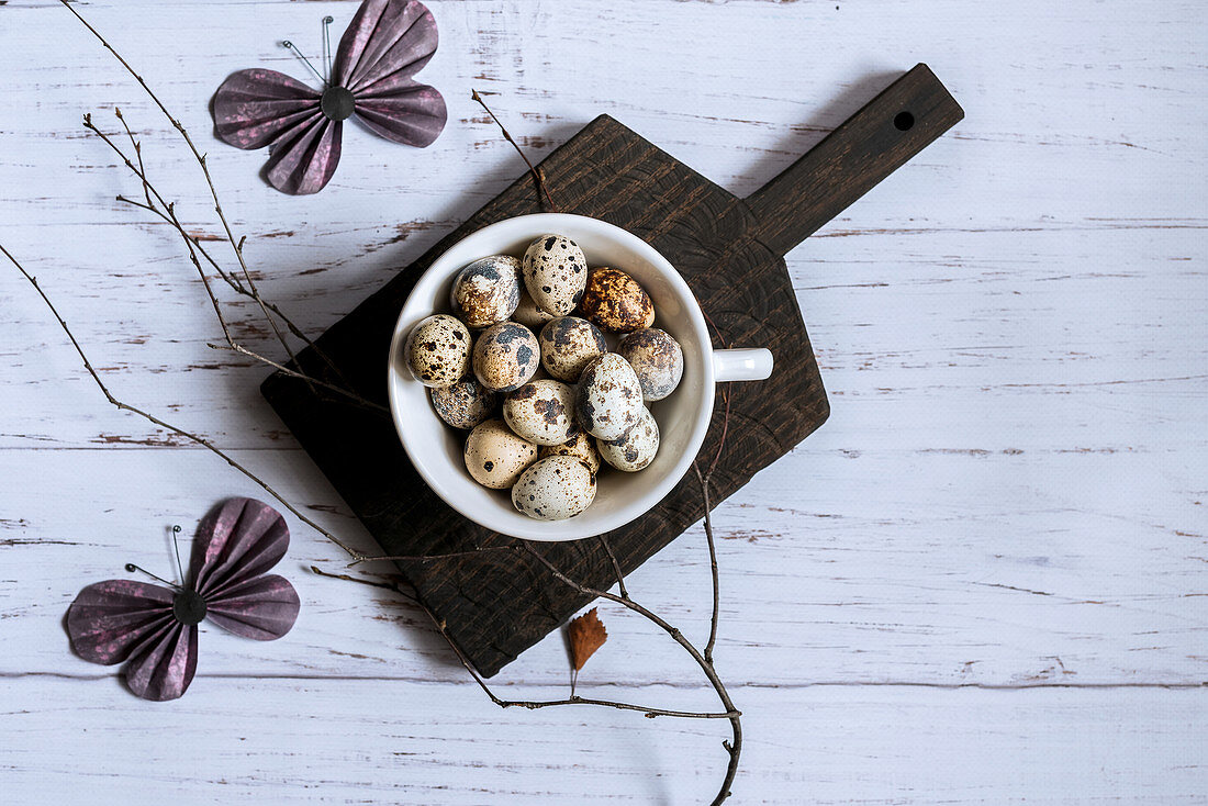 Top view of quail eggs in a white bowl on a black cutting board decorated with a bare tree branch and paper butterflies