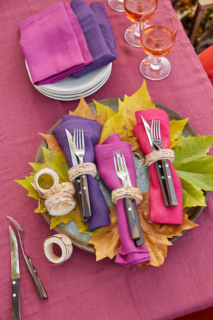 DIY napkin rings made from birch branches, colourful napkins and cutlery on autumnal leaves