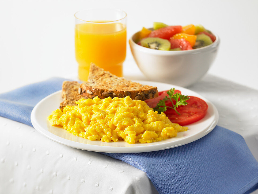 Scrambled eggs served with bread, tomatoes, orange juice and fruit salad
