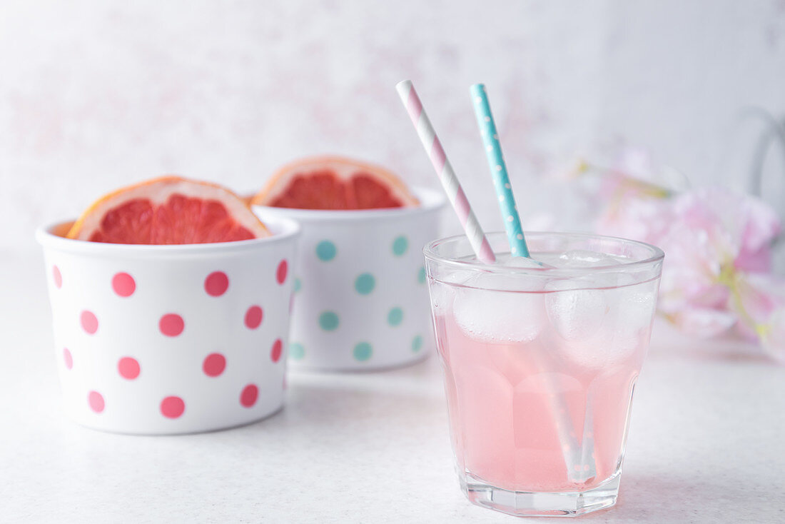 A glass of pink colored grapefruit infused water with pastel colored beverage straws, halved grapefruit in polka dotted bowls