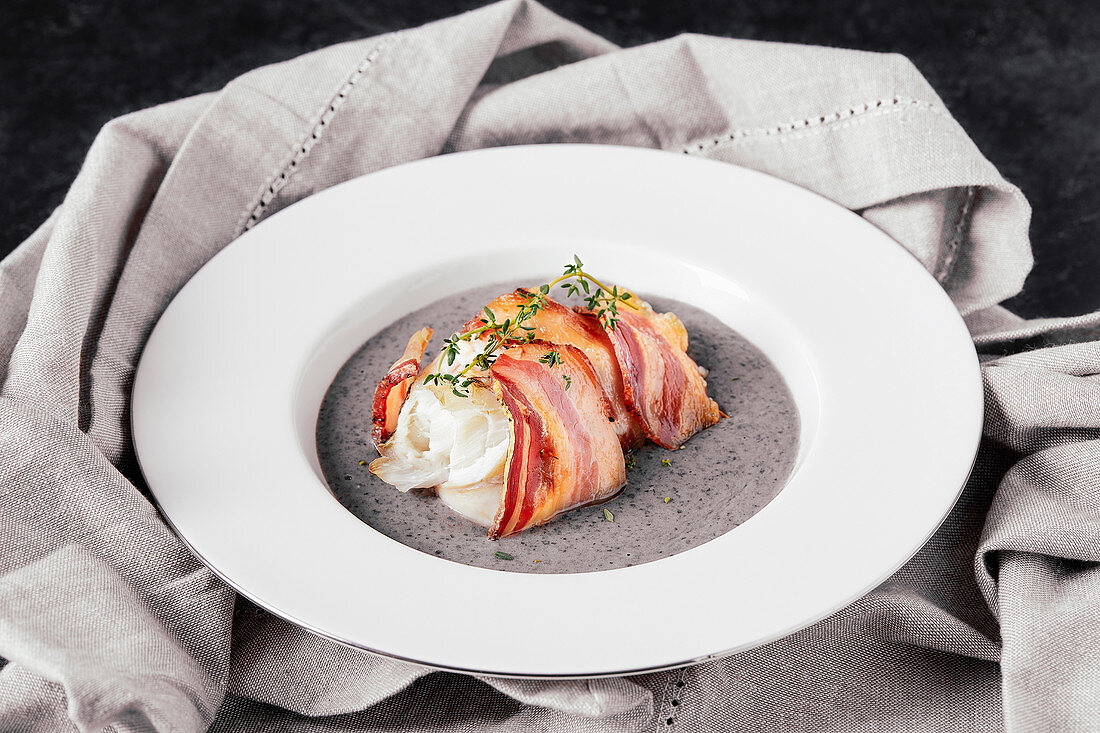 Plate with baked anglerfish fillet, wrapped in bacon, on black beans and potato cream