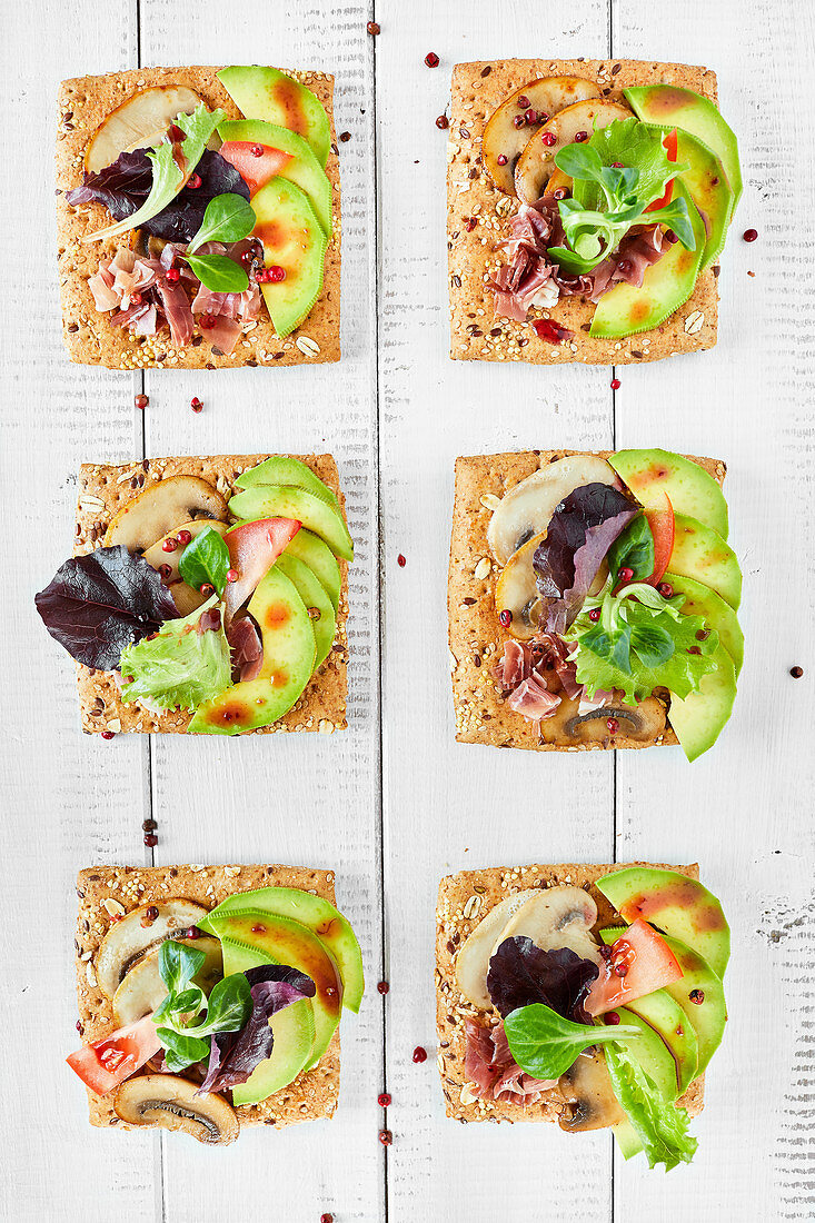 Canapes with mushrooms, avocado, salad, prosciutto on bread crouton with seeds, white wooden background