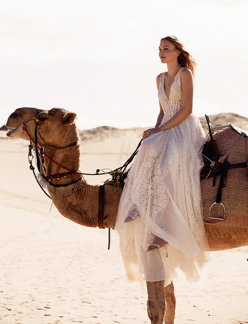 A young woman sitting on a camel wearing a long, white wedding dress