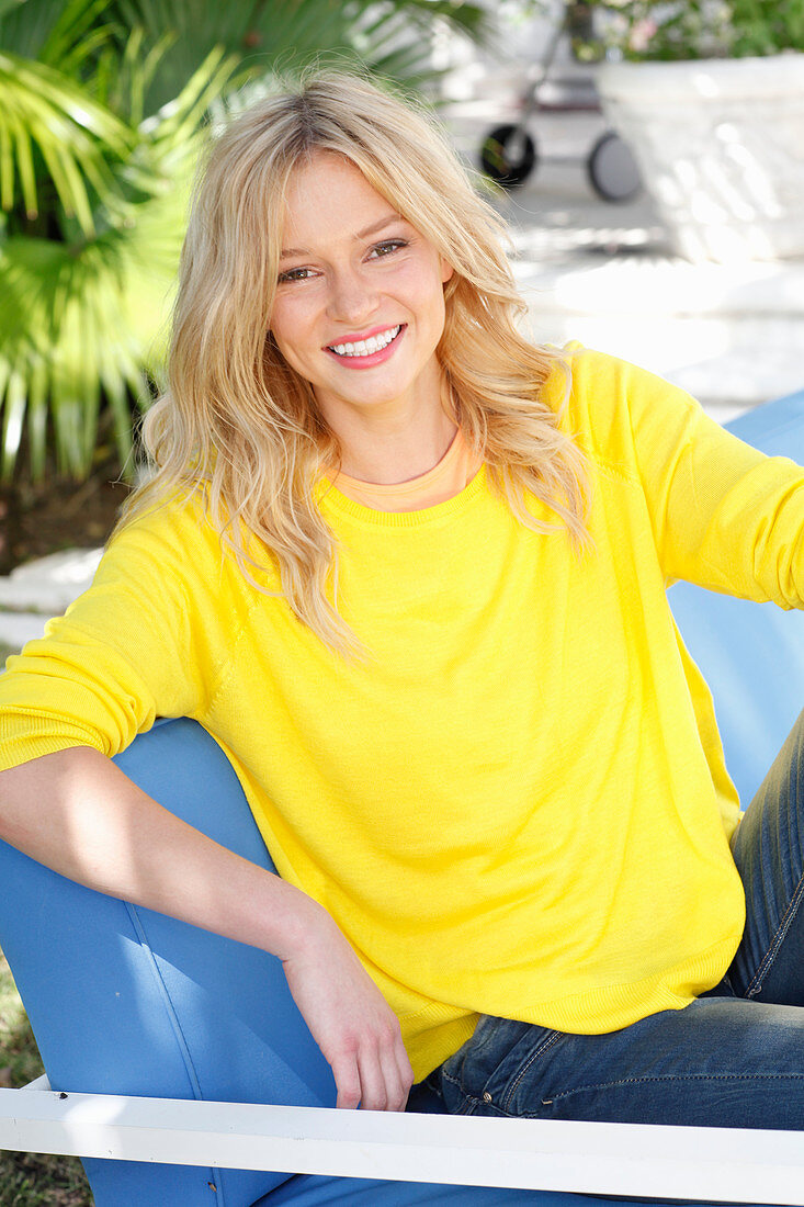 A young blonde woman wearing a yellow jumper sitting outside on a upholstered bench