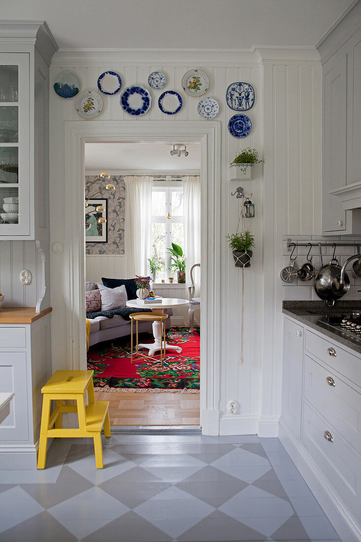 Yellow step stool in fitted kitchen with painted board floor, decorative wall plates on wood-panelled wall above door frame and view into living room