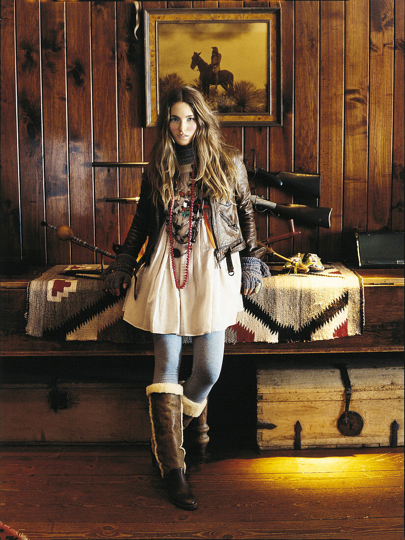 A young woman wearing a light dress, a leather jacket and boots in a hunting lodge
