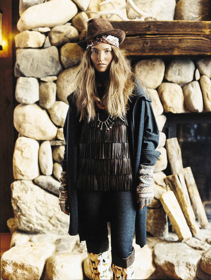 A young woman wearing a hat, a dark coat and leggings in front of a rustic, natural stone fireplace