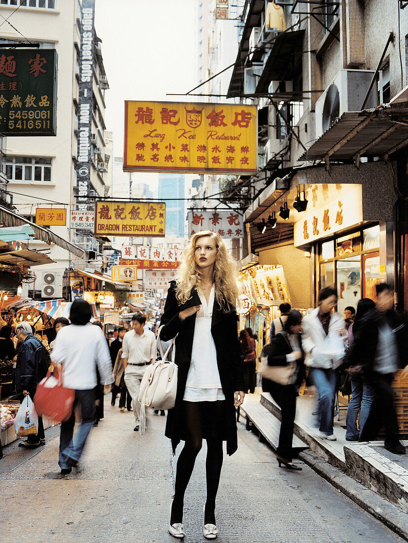 Blonde Frau in schwarz-weißem Kleid und schwarzem Mantel auf der Straße von Hong Kong