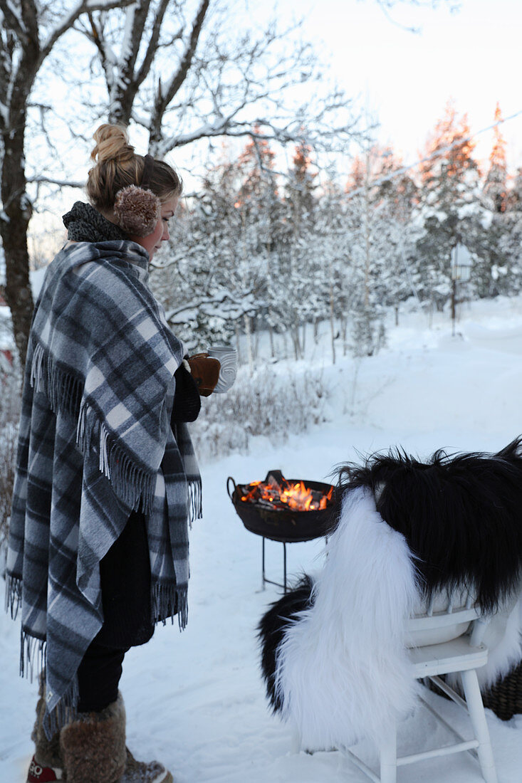 Woman standing next to char with fur blanket and fire bowl in snowy landscape