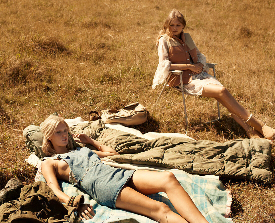 A blonde woman lying on a blanket and a brunette woman sitting on a camping chair in a meadow
