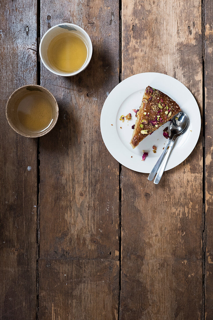 Stück Pistazienkuchen mit Rosenblättern und zwei Teetassen auf Holztisch