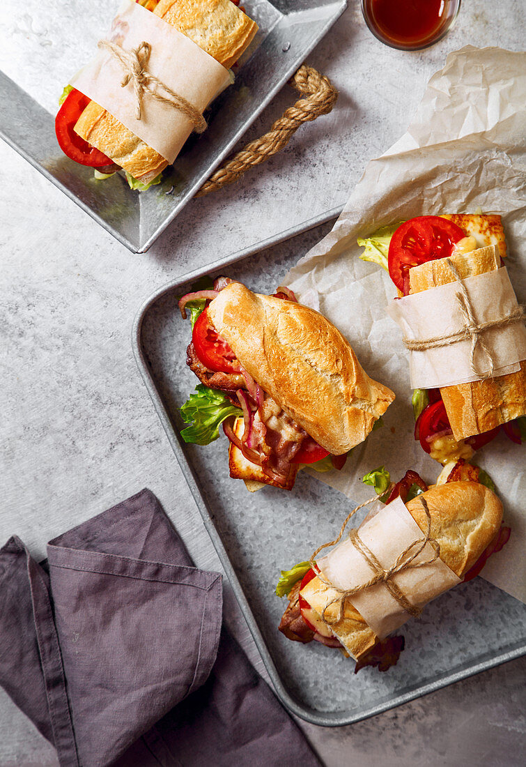Fresh baguette sandwich bahn-mi styled, bacon, roasted cheese, tomatoes and lettuce on metallic tray background