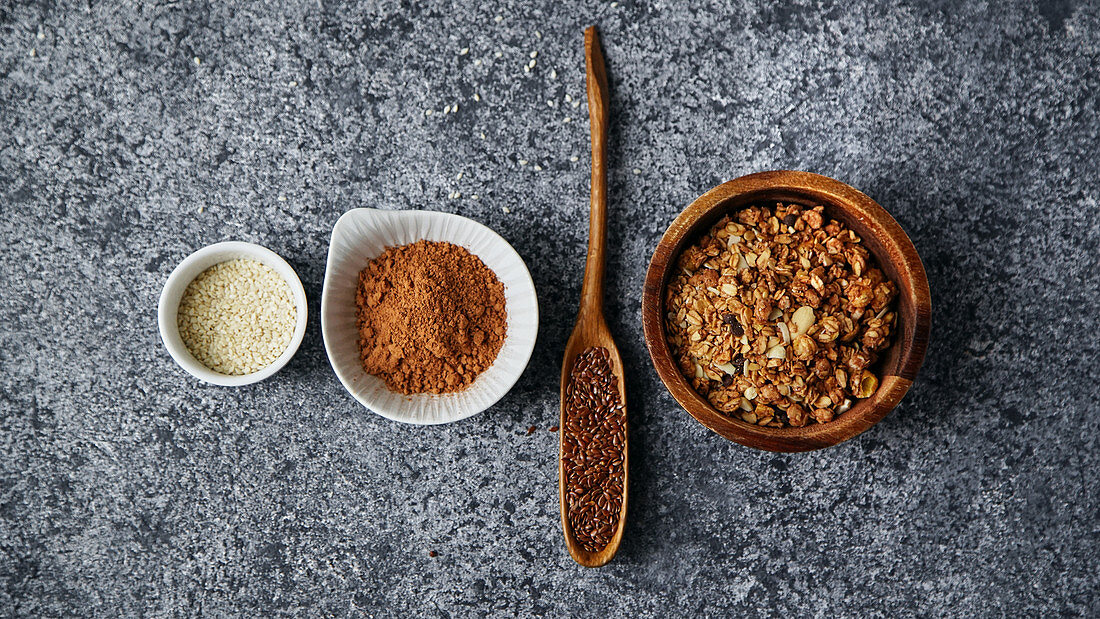 Healthy sweets cooking ingredients: granola, sesame and flax seeds, cocoa in separate bowls on gray background