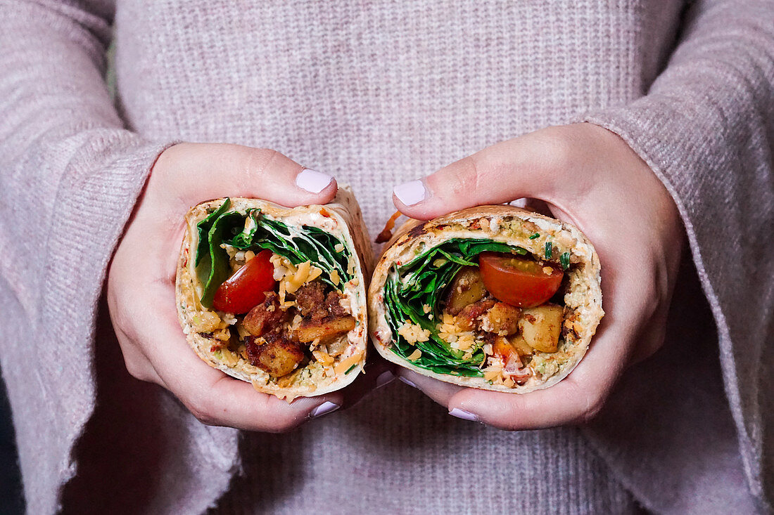 Frau hält Burrito mit gebratenen Kartoffelwürfeln, Tomaten, Frischkäse, Cheddar und Spinat